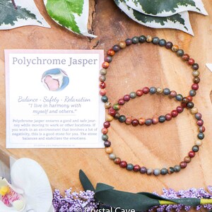 May include: A Polychrome Jasper bracelet featuring red, brown, and grey beads. The bracelet is displayed on a wooden surface with green leaves and a white flower. The accompanying card reads "Polychrome Jasper" and "Balance - Safety - Relaxation" with the quote "I live in harmony with myself and others."