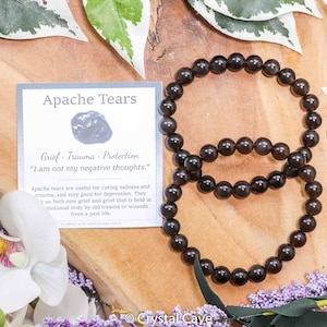 May include: Two black Apache Tears bracelets on a wooden surface. The bracelets are made of round beads and are stacked on top of each other. A small card with the text "Apache Tears" and "Grief - Trauma - Protection" is visible in the background.
