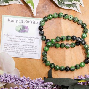 Pode incluir: Um bracelete de pedras preciosas verde e preto feito de contas de rubi em zoisita. O bracelete está sobre uma superfície de madeira com flores no fundo. O texto no cartão diz: "Força de vontade - Transformação - Espiritualidade. "O poder positivo que está em mim é mais forte do que meus medos."