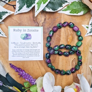 May include: A green and pink ruby in zoisite bracelet. The bracelet is made of smooth, rounded stones. The card reads "Ruby in Zoisite" and "Willpower - Transformation - Spirituality" and "The positive power that is in me is stronger than my fears."