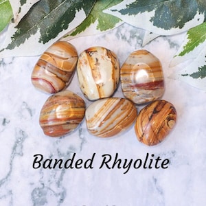 May include: A collection of six banded rhyolite tumbled stones. The stones are a mix of brown, white, and orange colors. The stones are arranged on a white marble surface. The text "Banded Rhyolite" is written in black below the stones. The text "Crystal Cave" is written in black below the text "Banded Rhyolite".