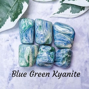 May include: Six tumbled blue green kyanite stones with white and green swirls. The stones are arranged in a circle on a light purple background. The text "Blue Green Kyanite" is written below the stones. The text "Crystal Cave" is written below the text "Blue Green Kyanite".