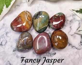 May include: Six smooth, polished, multi-colored jasper stones. The stones are various shades of red, brown, green, and white. The stones are arranged in a circle on a white marble surface. "Fancy Jasper" is written in black text below the stones. "Crystal Cave" is written in black text below the phrase "Fancy Jasper".