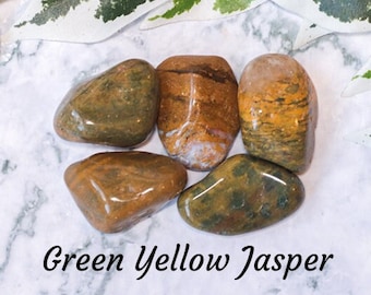 May include: Five tumbled green and yellow jasper stones on a white marble background. The stones are smooth and polished, with a variety of patterns and colors.