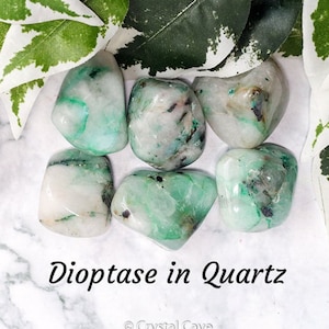 May include: A collection of six tumbled stones of dioptase in quartz. The stones are a mix of white and green, with the green being a bright, vibrant shade. The stones are smooth and polished, and they are arranged in a circle on a white marble surface.
