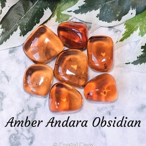Puede incluir: Una colección de siete piedras de obsidiana andara ámbar pulidas. Las piedras son lisas y pulidas, con un color translúcido similar al ámbar. Las piedras están dispuestas en un grupo sobre una superficie de mármol blanco.
