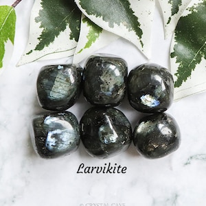 Pode incluir: Seis pedras lisas de Larvikite de cor cinza escuro com reflexos iridescentes azuis e verdes. As pedras estão dispostas em círculo sobre uma superfície branca.