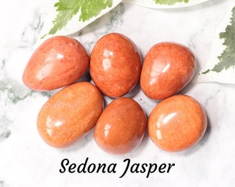 Op de afbeelding: Zes gladde, gepolijste, oranje-rode Sedona Jaspis stenen. De stenen zijn getrommeld en hebben een natuurlijke, aardse uitstraling. De tekst "Sedona Jasper" is in zwarte cursieve letters onder de stenen geschreven.