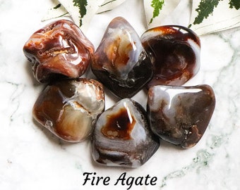 May include: A collection of six tumbled fire agate stones. The stones are a mix of brown, red, and white with a banded pattern. The stones are on a white marble surface.
