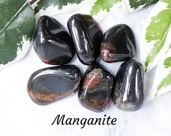 May include: A collection of six tumbled Manganite stones. The stones are black with red and silver streaks. The stones are arranged in a circle on a white surface.