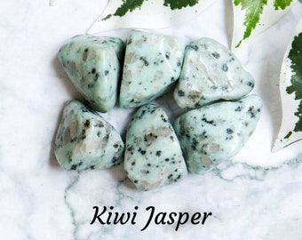 May include: A collection of six smooth, light green kiwi jasper tumbled stones with dark green speckles. The stones are arranged in a circle on a white marble surface.