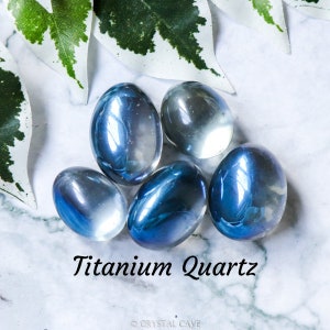 May include: Five smooth, oval-shaped titanium quartz crystals in shades of blue and white. The crystals are arranged in a cluster on a white marble surface.