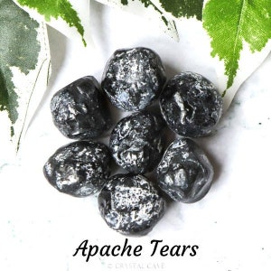 Könnte beinhalten: Sieben schwarze Apache Tears Steine, auch bekannt als Obsidian, mit einer rauen, strukturierten Oberfläche. Die Steine sind in einem Kreis auf einem weißen Hintergrund angeordnet.