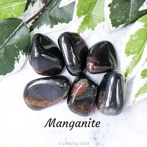 May include: A collection of six tumbled Manganite stones. The stones are black with red and silver streaks. The stones are arranged in a circle on a white surface.