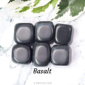 Könnte beinhalten: Sechs glatte, dunkelgraue Basalt-Trommelsteine, jeder etwa so groß wie eine 20-Cent-Münze, in einem quadratischen Muster auf einer weißen Marmorfläche angeordnet. Das Wort "Basalt" ist in schwarzer Kursivschrift unter den Steinen geschrieben.