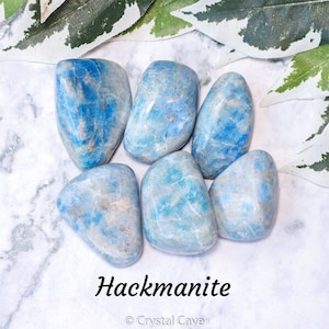 May include: A collection of six smooth, light blue Hackmanite tumbled stones. The stones have a mottled, cloudy appearance with a hint of white. The stones are arranged in a circle on a white marble surface. The text "Hackmanite" and "Crystal Cave" are printed below the stones.