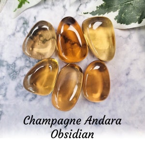 Puede incluir: Seis piedras lisas y pulidas de color ámbar dorado, conocidas como Obsidiana Andara Champagne, están dispuestas sobre una superficie de mármol blanco. Las piedras son de diversas formas y tamaños, algunas con un ligero tono ahumado.