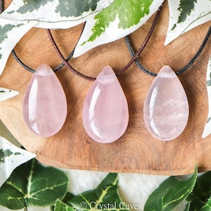Op de afbeelding: Drie roze rozenkwarts druppel hangers aan bruine leren koorden. De hangers worden getoond op een houten oppervlak met groene bladeren.