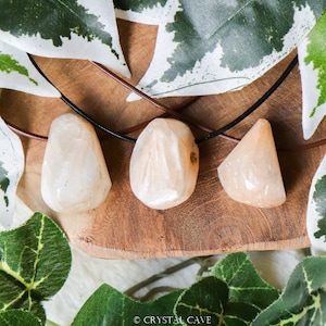 May include: Three light orange and white gemstone pendants on a black cord necklace. The pendants are on a wooden surface with green leaves in the background.