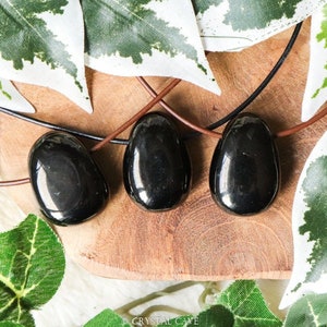 Op de afbeelding: Drie zwarte obsidiaan hangers op bruine leren koorden, gepresenteerd op een houten oppervlak met groene bladeren.