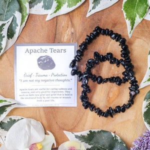 May include: Two black Apache Tears chip bead bracelets are displayed on a wooden surface, next to a card with the text "Apache Tears". The card also includes the phrases "Grief, Trauma, Protection" and "I am not my negative thoughts."