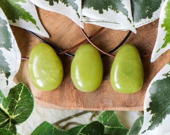 May include: Three green jade stone pendants on a brown leather cord. The stones are smooth and polished, and they are surrounded by green leaves.
