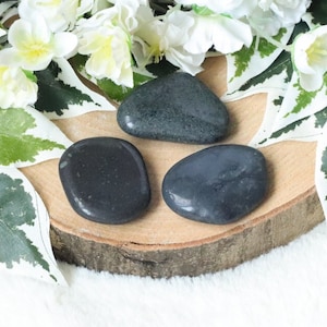 May include: Three smooth, dark gray tumbled stones on a wooden surface. The stones are oval shaped and have a polished finish.