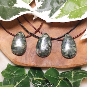 May include: Three teardrop-shaped pendants with dark green and speckled patterns, each suspended on a leather cord. The pendants are displayed on a wooden surface, with green and white leaves in the background. The pendants are a natural stone.