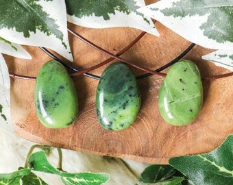 May include: Three green jade stone pendants on a brown leather cord. The stones are smooth and polished, with a light green color and dark green veins. The stones are displayed on a wooden surface with green leaves.