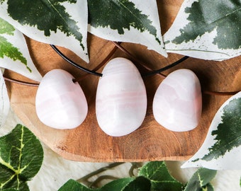 May include: Three pink, smooth, oval-shaped stones on a wooden surface with green leaves. The stones are connected by a brown leather cord.