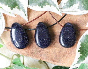 May include: Three blue sunstone pendants on a wooden surface with green leaves in the background. The pendants are smooth and have a sparkly texture.