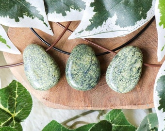 May include: Three green and white serpentine stone pendants on a brown leather cord. The stones are irregularly shaped and have a mottled pattern.
