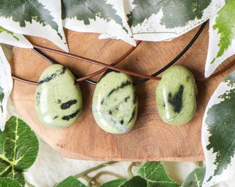 May include: Three green and black gemstone pendants on brown leather cords. The stones are smooth and polished, with a teardrop shape. The pendants are displayed on a wooden surface with green leaves.