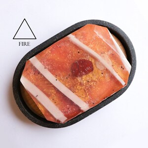 May include: A red, orange, and gold soap bar with white stripes and gold flecks, resting in a black oval dish with a wooden base. The soap is labeled "FIRE" with a triangle symbol.
