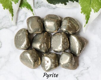 May include: A group of eight smooth, metallic, gray pyrite tumbled stones on a white marble surface. The stones are arranged in a circle with the word "Pyrite" written in black below them. The text "© CRYSTAL CAVE" is written in black below the word "Pyrite".