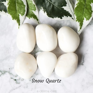 May include: Six smooth, white snow quartz tumbled stones on a marble surface. The stones are oval shaped and have a polished finish. The text "Snow Quartz" is written below the stones.