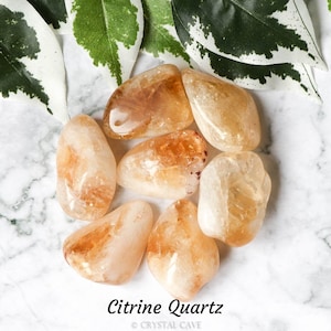 May include: A collection of seven tumbled citrine quartz stones, ranging in size and shape, with a light yellow to orange hue. The stones are arranged in a circle on a white marble surface. Citrine Quartz.