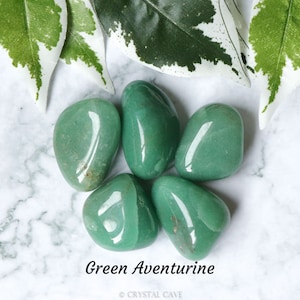 May include: Five smooth, green aventurine tumbled stones on a white marble background. The stones are a vibrant green color and have a glossy finish.