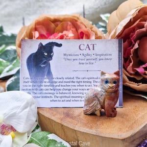 Könnte beinhalten: Eine kleine, geschnitzte Katzenfigur aus Stein mit blauen Augen steht neben einer Karte mit dem Wort "CAT" und Text über Mystik, Beweglichkeit und Inspiration. Die Figur ist eine Mischung aus Orange, Braun und Weiß. Der Hintergrund umfasst Blumen.