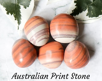 Op de afbeelding: Vijf gladde, afgeronde Australische Print Stones, elk met unieke, draaiende patronen in oranje, bruin en wit. De stenen zijn in een cluster op een wit marmeren oppervlak gerangschikt.