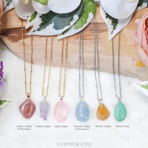 May include: Seven necklaces with different colored gemstone pendants. The necklaces are on a wooden surface with flowers in the background. The necklaces are available in gold and silver chains with 1.5mm and 2mm sizes. The text on the image reads: "GOLD 1.5MM WITH BALLS", "GOLD 1.5MM", "GOLD 2MM", "SILVER 1.5MM WITH BALLS", "SILVER 1.5MM", "SILVER 2MM".