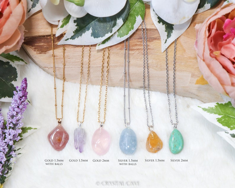 May include: Seven necklaces with different colored gemstone pendants. The necklaces are on a wooden surface with flowers in the background. The necklaces are available in gold and silver chains with 1.5mm and 2mm sizes. The text on the image reads: "GOLD 1.5MM WITH BALLS", "GOLD 1.5MM", "GOLD 2MM", "SILVER 1.5MM WITH BALLS", "SILVER 1.5MM", "SILVER 2MM".