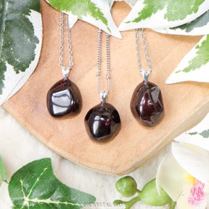 May include: Three silver necklaces with dark red gemstone pendants. The pendants are smooth and irregularly shaped, resembling small pebbles. The necklaces are displayed on a wooden surface.