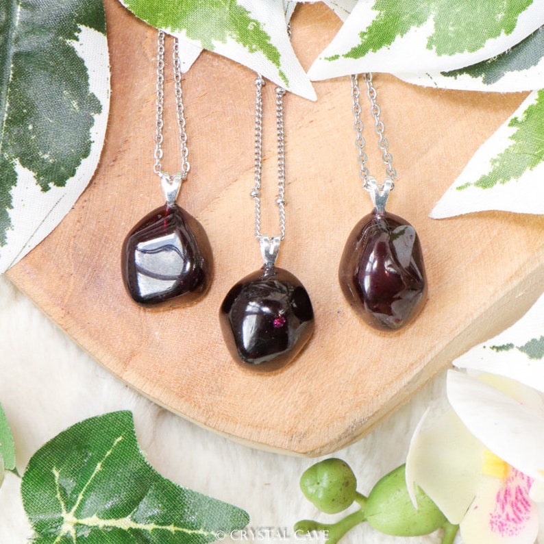 May include: Three silver necklaces with dark red gemstone pendants. The pendants are smooth and irregularly shaped, resembling small pebbles. The necklaces are displayed on a wooden surface.