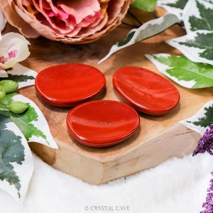 Può includere: Tre pietre di diaspro rosso a forma di palma su una superficie di legno. Le pietre sono lisce e levigate, e hanno un aspetto naturale e terroso.