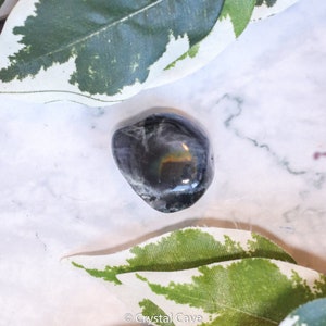 May include: A smooth, black, tumbled obsidian stone with iridescent rainbow flashes. The stone is resting on a white marble surface with green leaves.