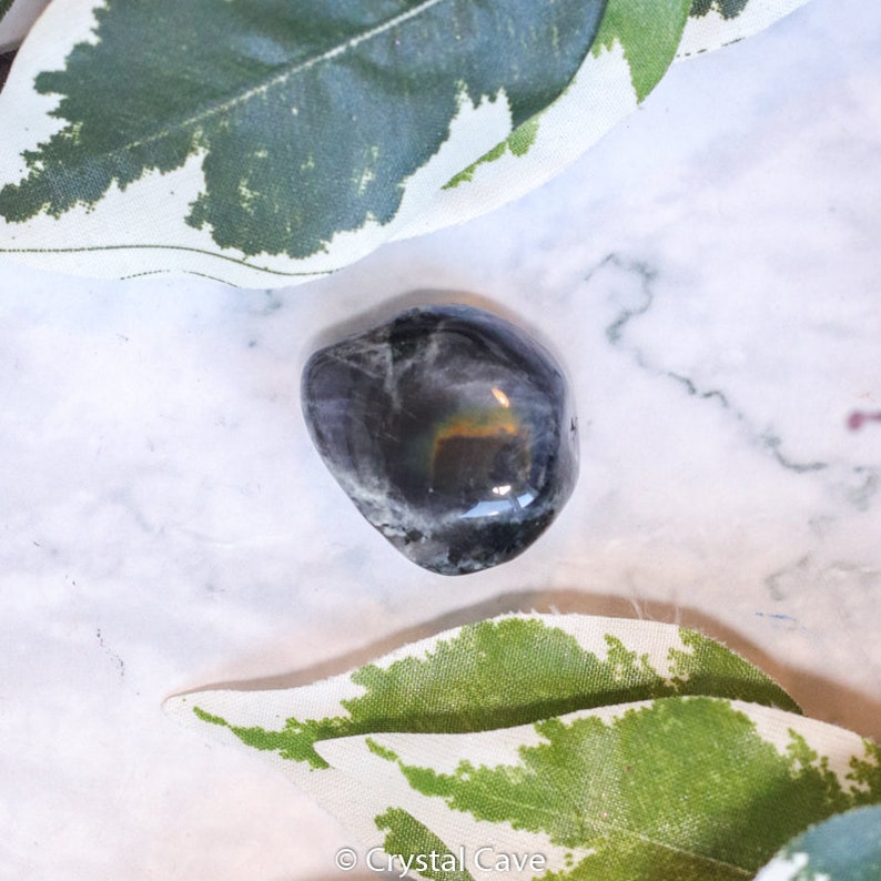 May include: A smooth, black, tumbled obsidian stone with iridescent rainbow flashes. The stone is resting on a white marble surface with green leaves.