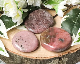 May include: Three pink and brown polished Rhodonite stones, also known as the love stone, on a wooden surface.