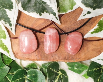 May include: Three pink rose quartz stones on a black leather cord necklace. The stones are smooth and polished, and they are arranged in a row on a wooden surface.