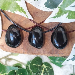 May include: Three black obsidian gemstone pendants on black leather cords. The pendants are smooth and polished, and they are arranged in a row on a wooden surface.